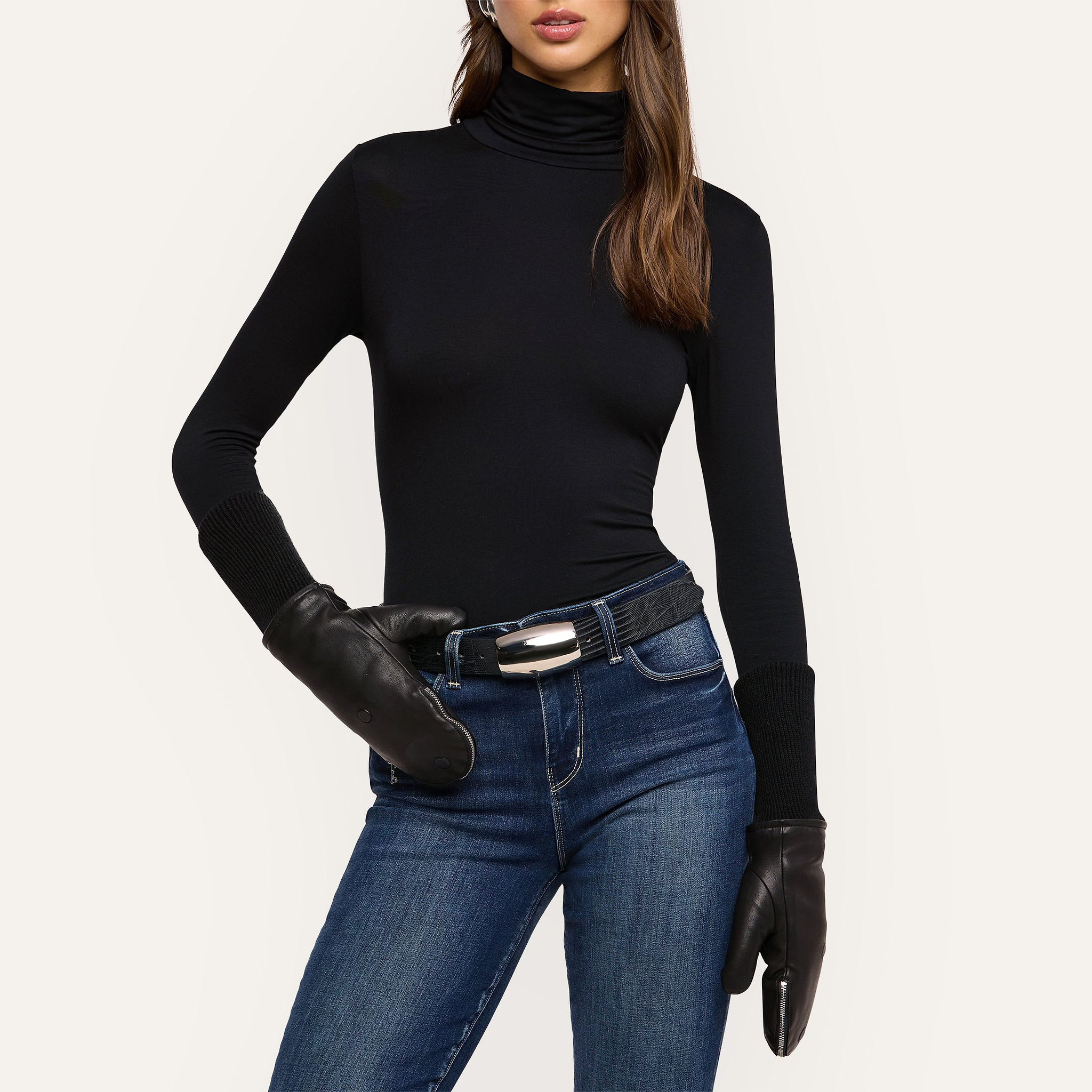A woman stands with one hand on her hip against a plain background, wearing a black turtleneck, blue jeans, a black belt, and the Tempo Mittens. Her face is partially visible.