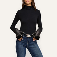 A woman wears a black turtleneck, blue jeans, and the Tempo Mittens luxury gloves, with her hands resting on her belt. The background is plain and light-colored.