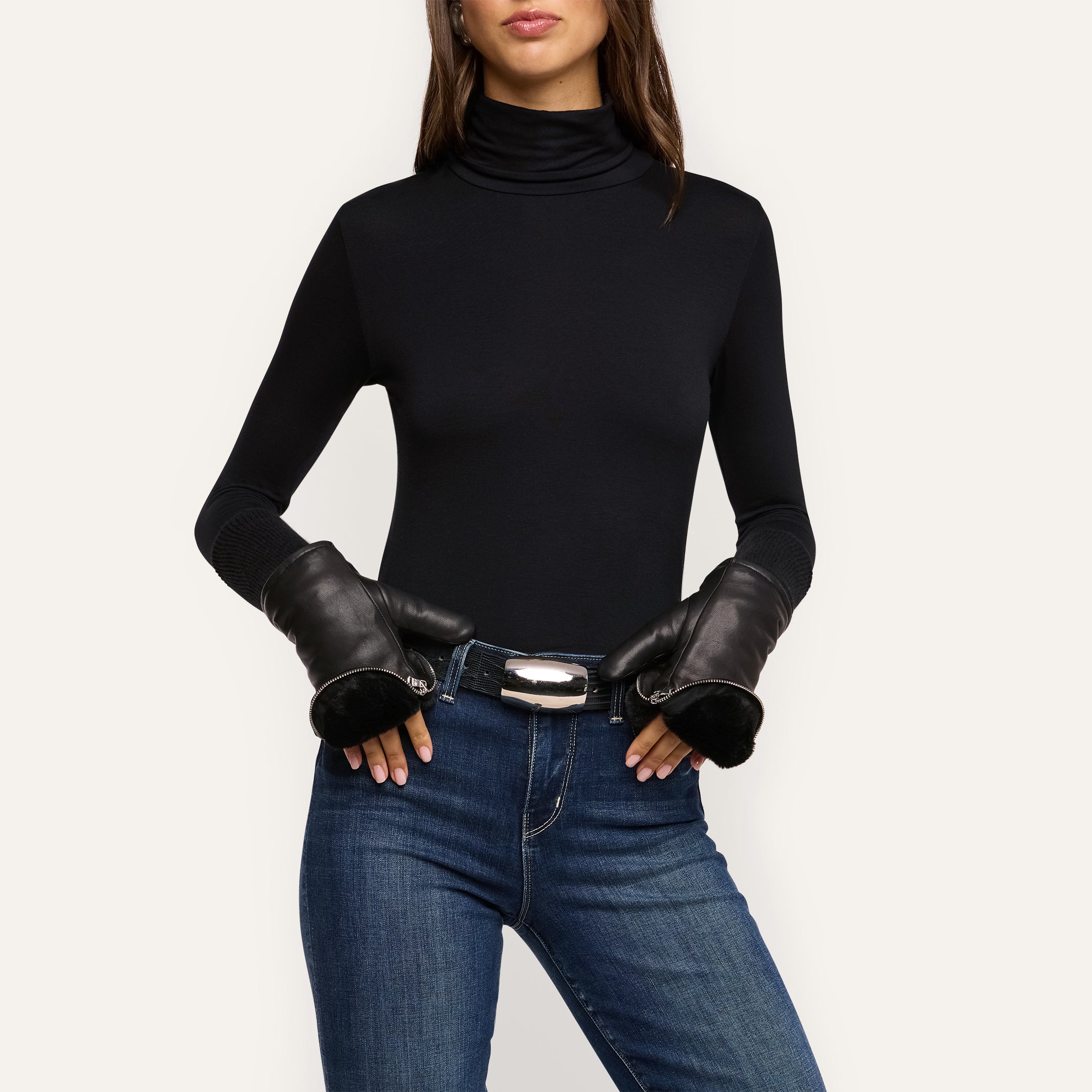 A woman wears a black turtleneck, blue jeans, and the Tempo Mittens luxury gloves, with her hands resting on her belt. The background is plain and light-colored.