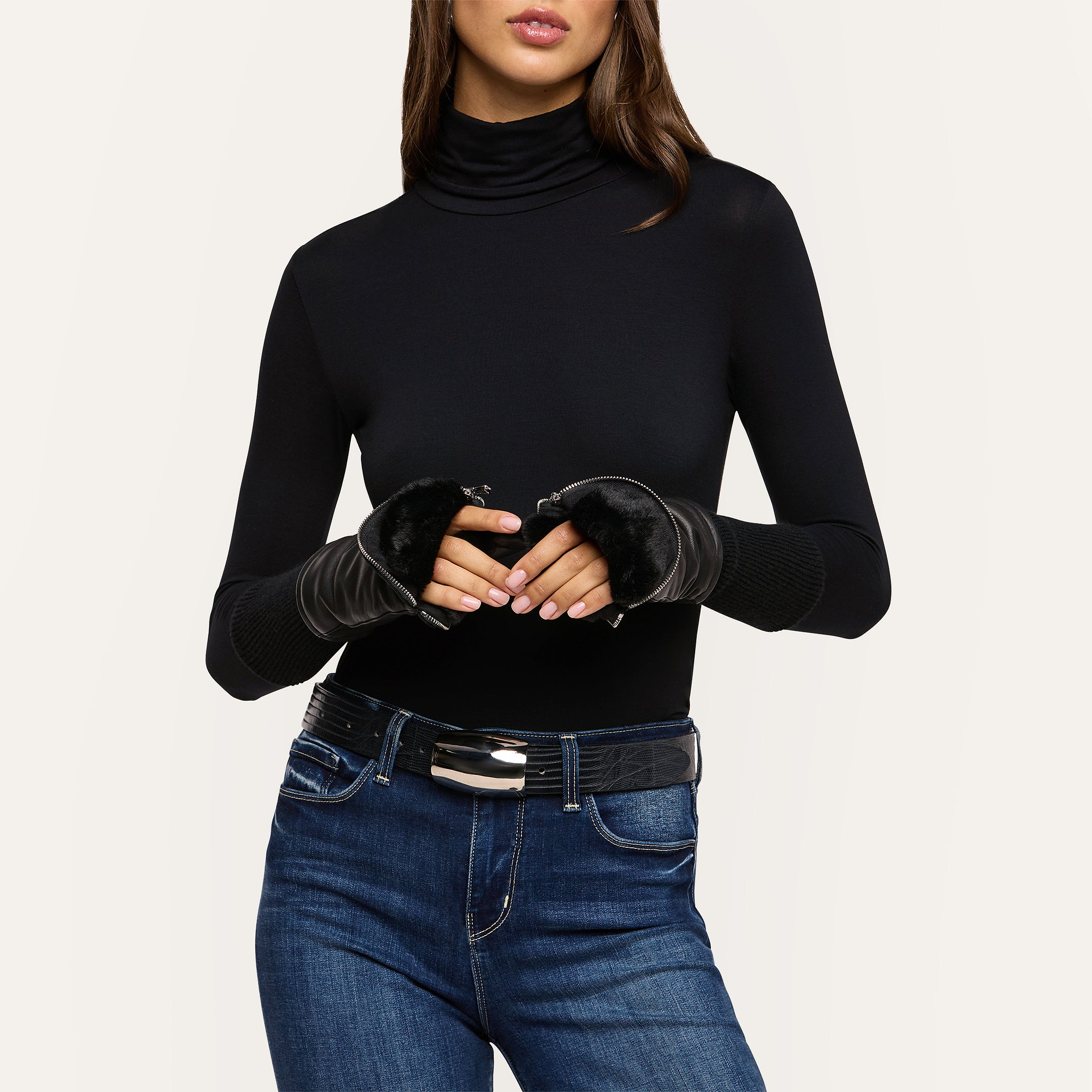 A woman poses against a light background with her hands together, wearing a black turtleneck, blue jeans, a black belt, and the Tempo Mittens—faux fur lined luxury black fingerless gloves.