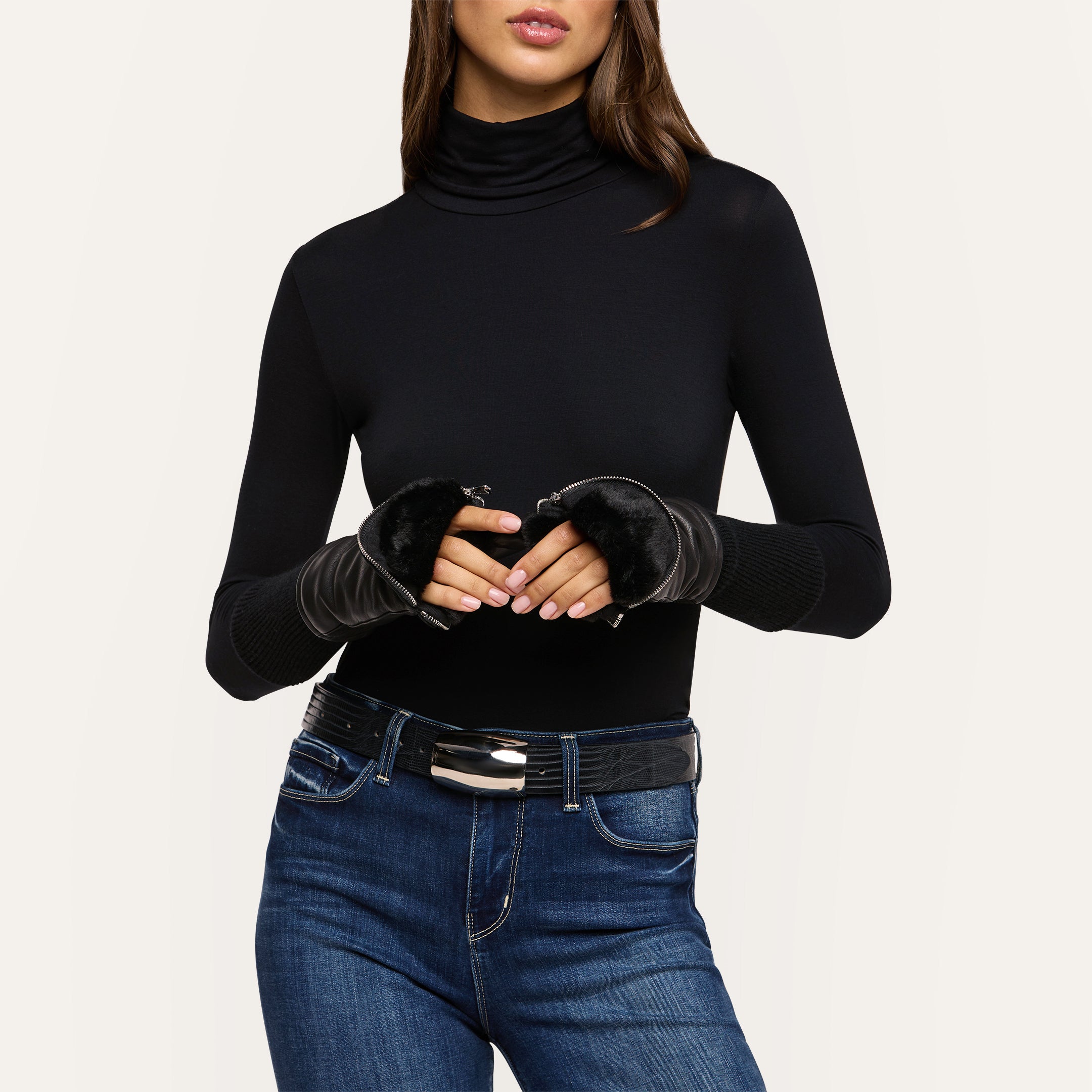 A woman poses against a light background with her hands together, wearing a black turtleneck, blue jeans, a black belt, and the Tempo Mittens—faux fur lined luxury black fingerless gloves.