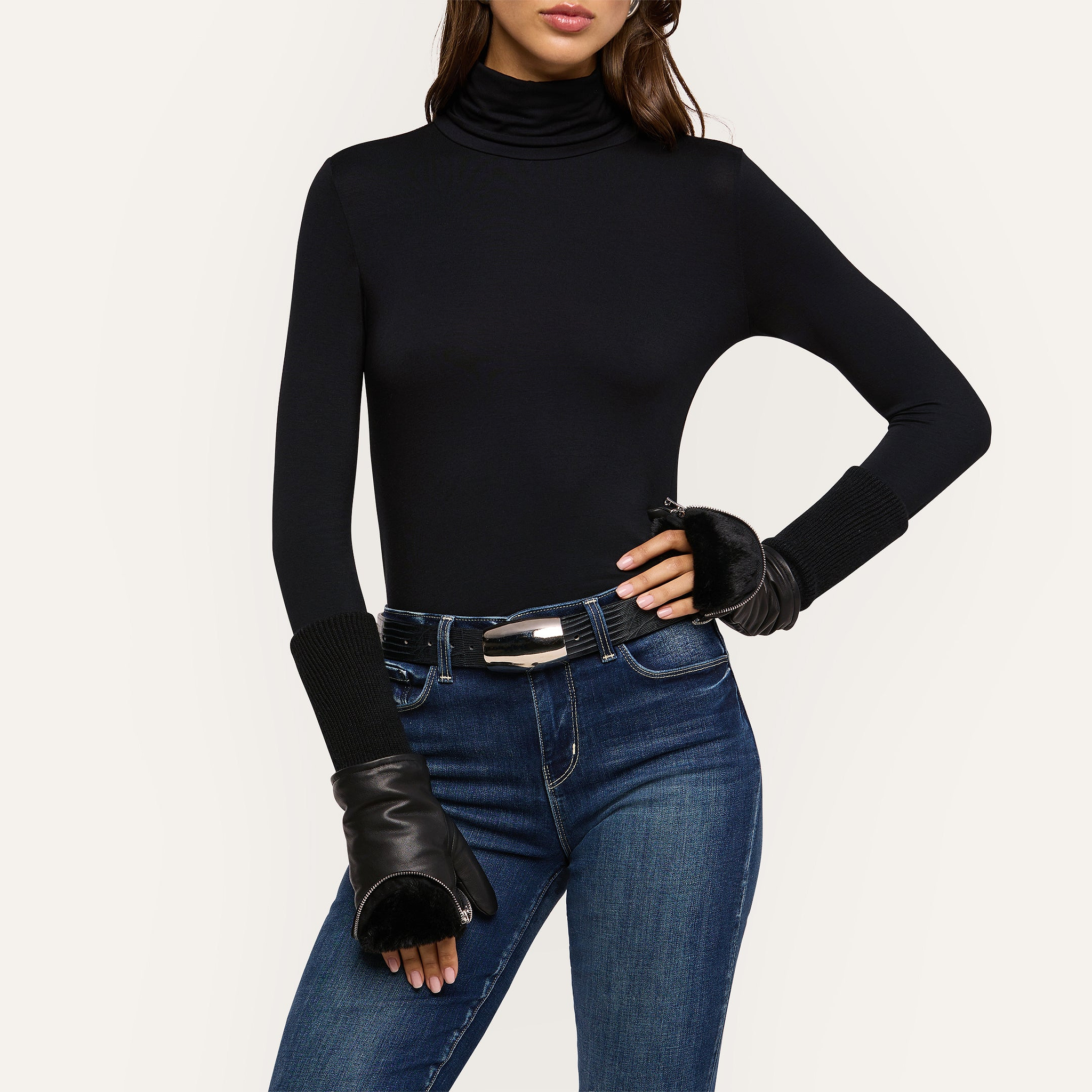 A woman poses against a plain background with one hand on her hip, dressed in a black turtleneck, dark blue jeans cinched with a silver-buckled black belt, and wearing the Tempo Mittens—luxurious black leather fingerless gloves featuring faux fur-lined trim and a stylish silver zipper detail.