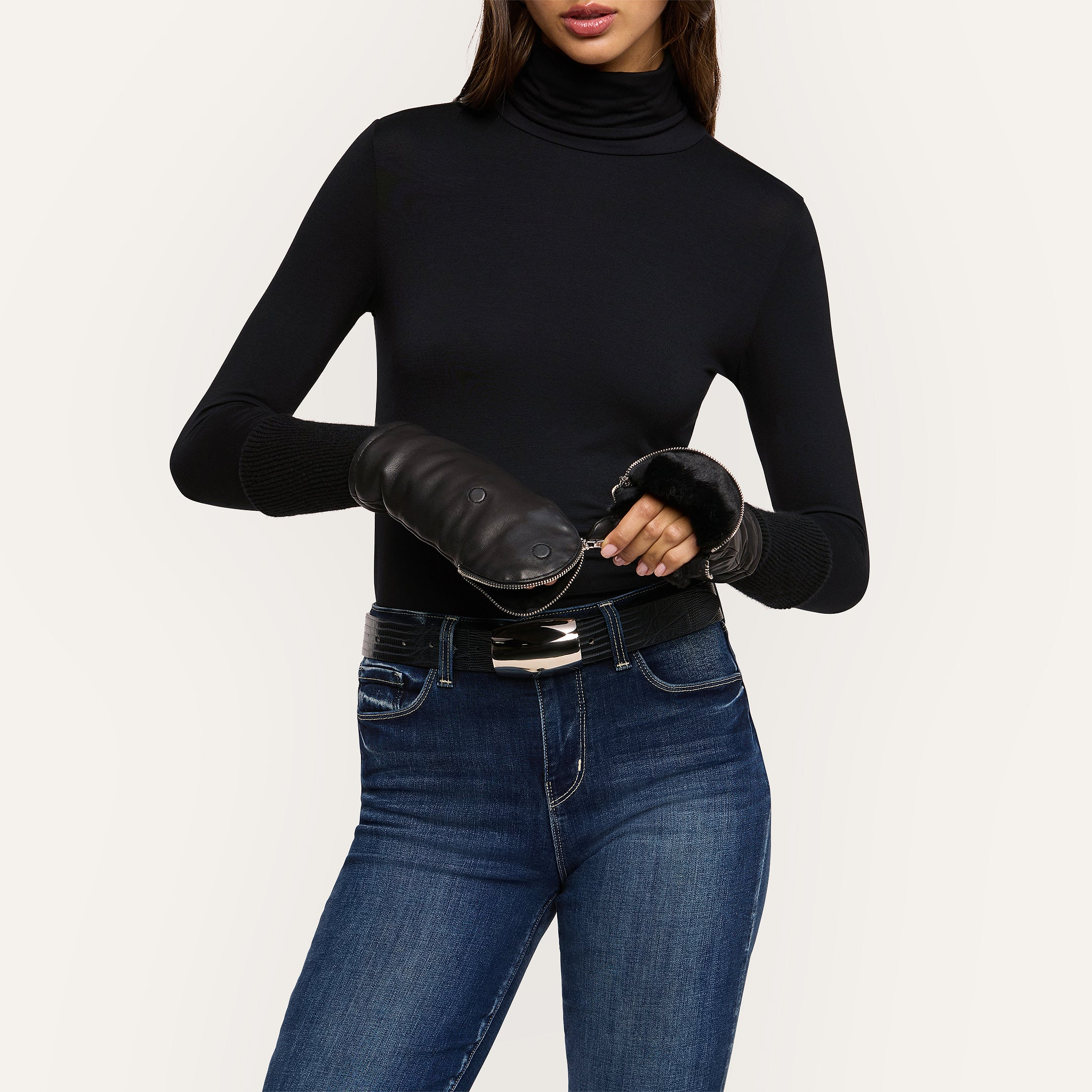 A woman stands against a plain light background, wearing a black turtleneck, blue jeans, and the Tempo Mittens—luxury gloves featuring a metallic buckle belt. Her face is partially out of frame.