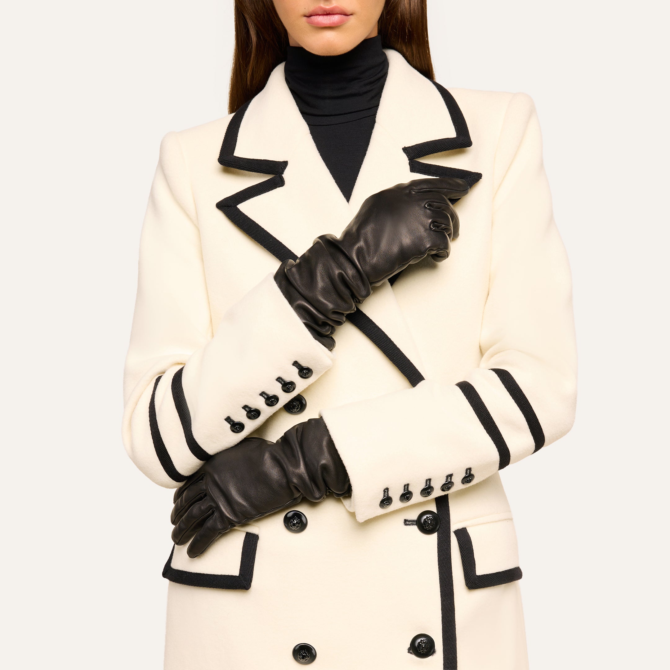 Wearing elegant Ballad Gloves made of lambskin leather, this person is dressed in a white coat with black trim and black buttons over a black turtleneck. Their arms are crossed, hands resting on opposite sleeves, and their face is partially out of frame.