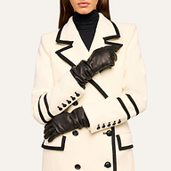 Wearing elegant Ballad Gloves made of lambskin leather, this person is dressed in a white coat with black trim and black buttons over a black turtleneck. Their arms are crossed, hands resting on opposite sleeves, and their face is partially out of frame.