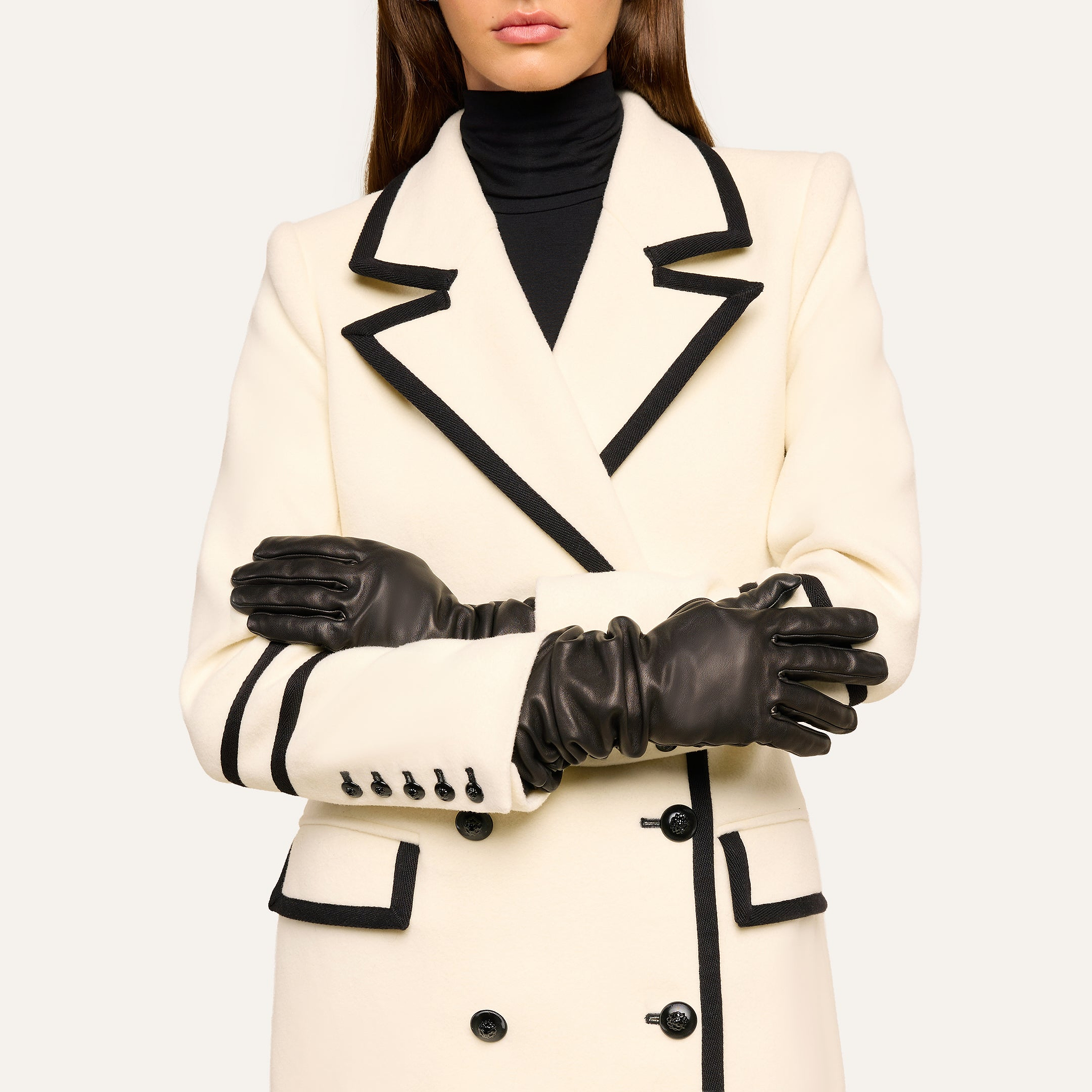 Woman wearing a white coat with black trim and black buttons, a black turtleneck, and the Ballad Gloves—long luxury lambskin leather gloves—with her arms crossed in front of her chest. Her face is partially out of the frame.