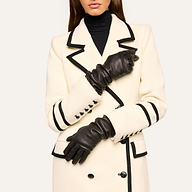 A woman stands with her arms crossed at her waist, wearing a white coat with black trim and black buttons, a black turtleneck, and the Ballad Gloves—luxurious lambskin leather gloves. Her face is partially out of frame.