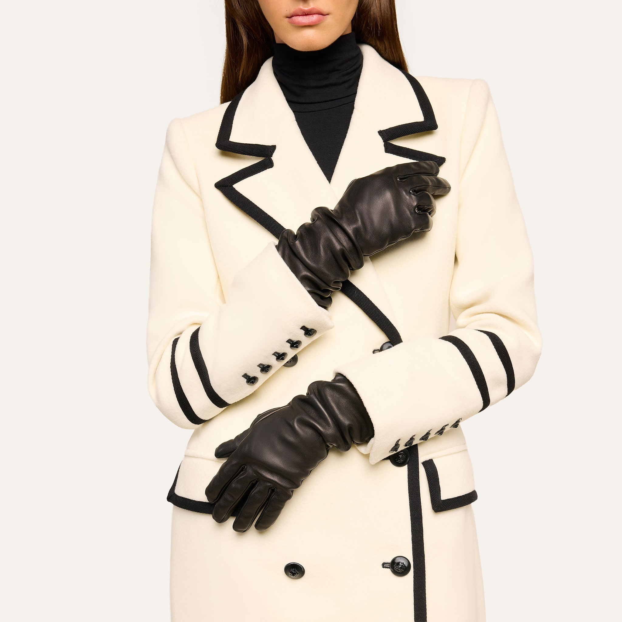 A woman stands with her arms crossed at her waist, wearing a white coat with black trim and black buttons, a black turtleneck, and the Ballad Gloves—luxurious lambskin leather gloves. Her face is partially out of frame.