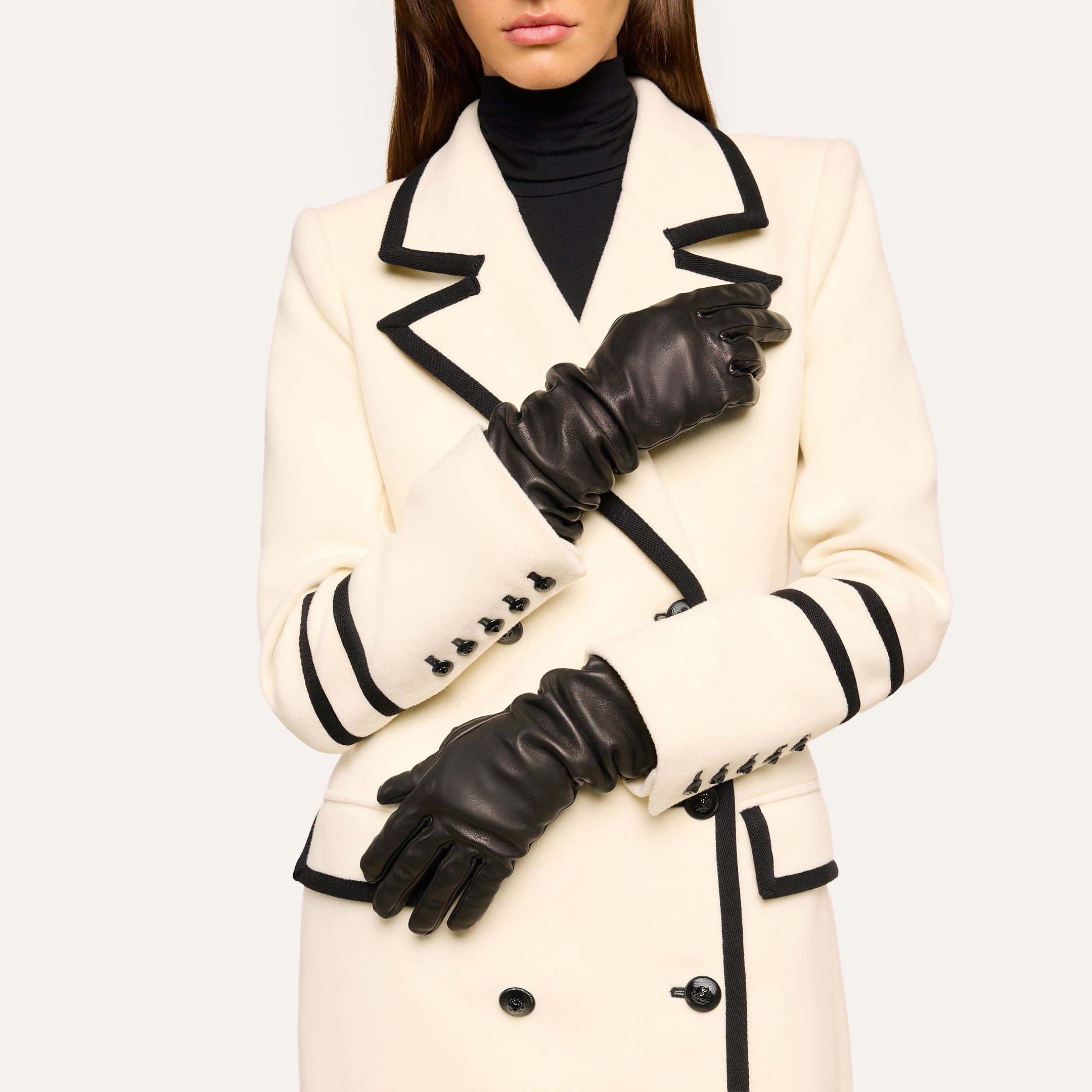 A woman stands with her arms crossed at her waist, wearing a white coat with black trim and black buttons, a black turtleneck, and the Ballad Gloves—luxurious lambskin leather gloves. Her face is partially out of frame.