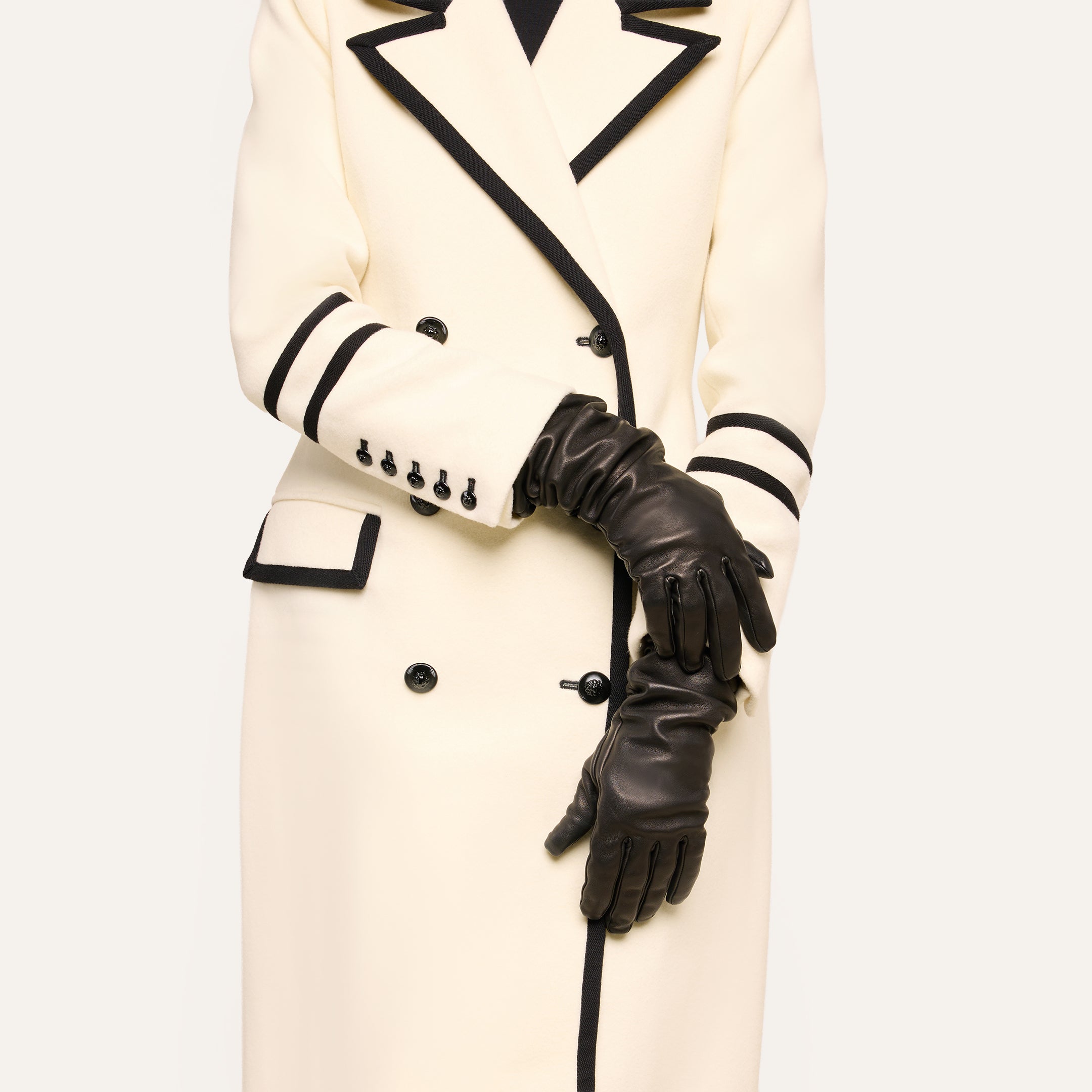 Model wears Ballad Gloves, long black lambskin leather gloves, paired with a cream-colored coat featuring black trim and buttons. The arms are crossed at the waist; face is not visible.