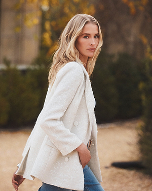 A woman with long blonde hair, wearing a textured white blazer and blue jeans, stands outdoors on a path with greenery and blurred autumnal trees in the background. She looks confidently toward the camera.
