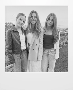 Three women stand close together outdoors, smiling at the camera. They are dressed casually; two wear jeans, and the one in the middle wears a long coat. Trees and a landscape are visible in the background. The image is in black and white.