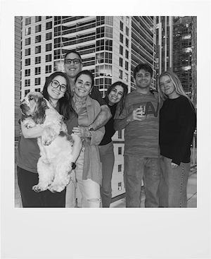 Six people and a dog pose and smile together on a balcony in a city. Tall modern buildings are in the background. The group appears cheerful, and one person is holding the dog. The image is in black and white.