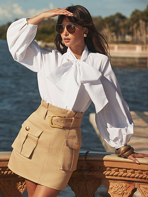 A woman wearing sunglasses, a white blouse with a large bow, and a beige skirt stands by a waterfront, shielding her eyes from the sun with one hand. Trees and a stone railing are visible in the background.