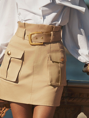 A person wearing the Maelis Mini Skirt with a high-rise waist, large front cargo pockets, gold buttons, and a wide matching belt, paired with a white blouse while standing outdoors.