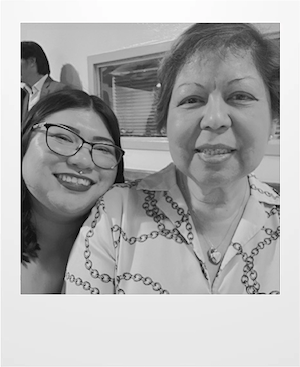 Two women smile for a close-up photo indoors. The woman on the left wears glasses and a dark top, while the woman on the right wears a patterned blouse and necklace. The image is in black and white.