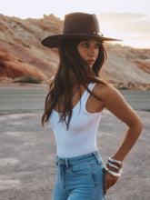 A woman stands outdoors near rocky terrain, wearing the Estella Fedora—a wide-brimmed espresso straw hat with a leather band—paired with a white sleeveless top and blue jeans, looking to the side with one hand on her hip.