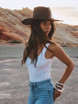 A woman wearing the Estella Fedora, featuring an espresso shade and leather band horse bit, pairs it with a white tank top and blue jeans as she stands outdoors in rocky, desert-like terrain, looking to the side with one hand on her hip.