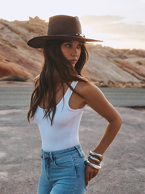 A woman wearing the L'AGENCE Estella Fedora stands on a rocky landscape at sunset, gazing to the side with her hand on her hip.