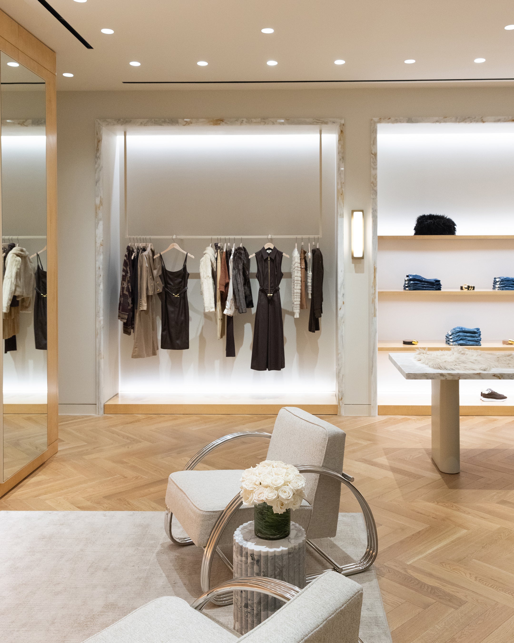 A boutique interior featuring a rack of dresses and jackets, folded denim on shelves, and two armchairs facing a table with a bouquet of white roses.