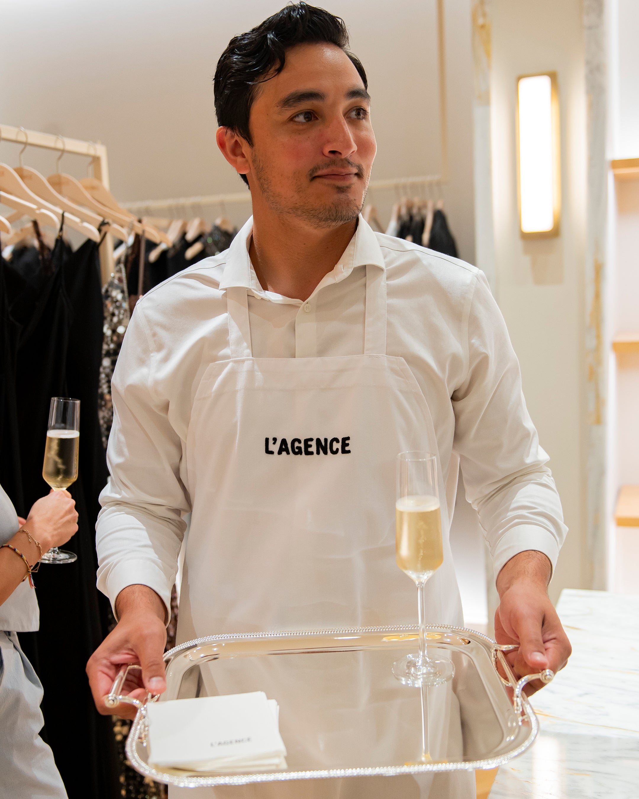 A man wearing a white LAGENCE apron holds a silver tray with a flute of champagne inside a clothing boutique.