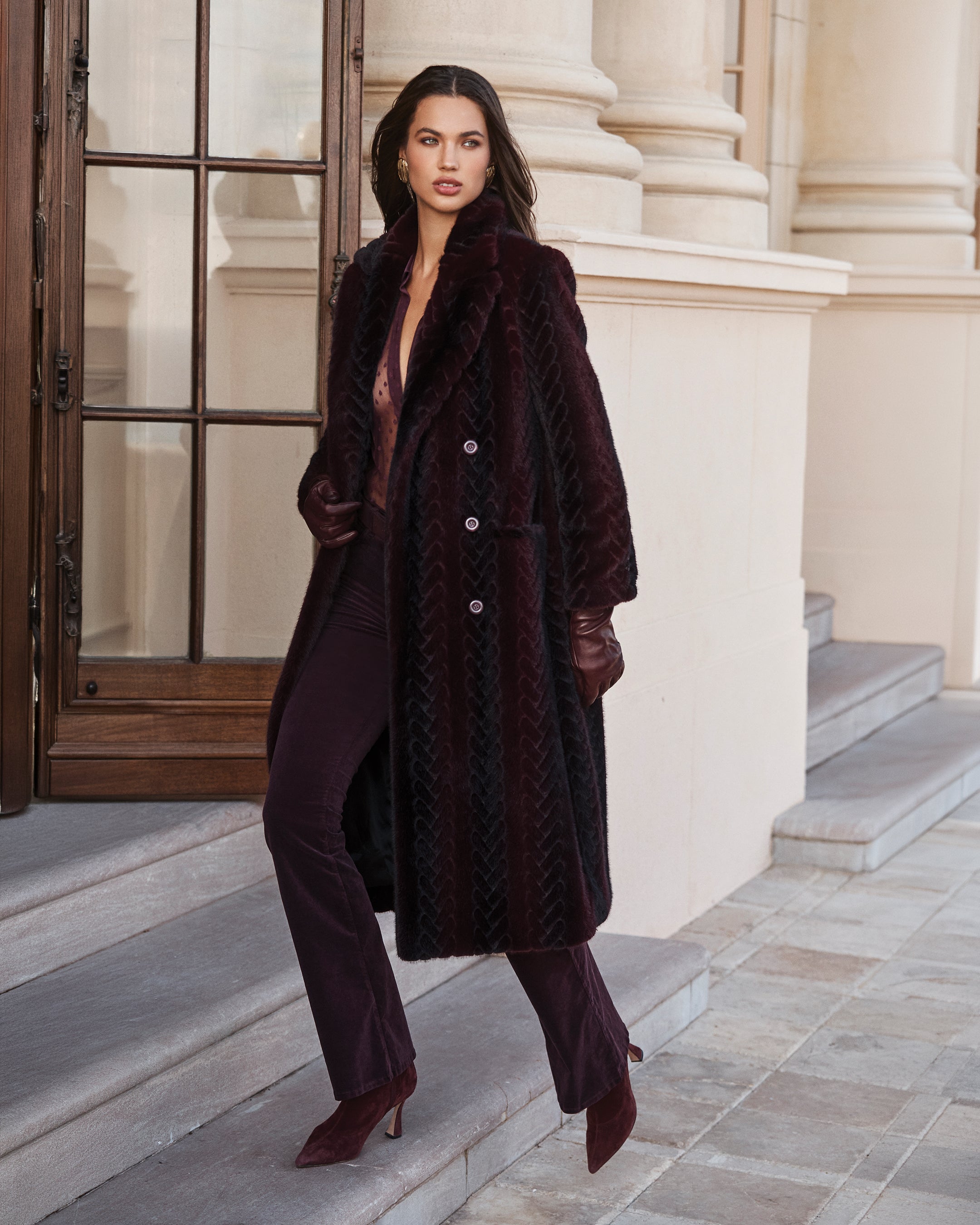 A woman in a long, dark textured coat, matching pants, gloves, and heels walks down stone steps outside a grand building with columns and large windows.