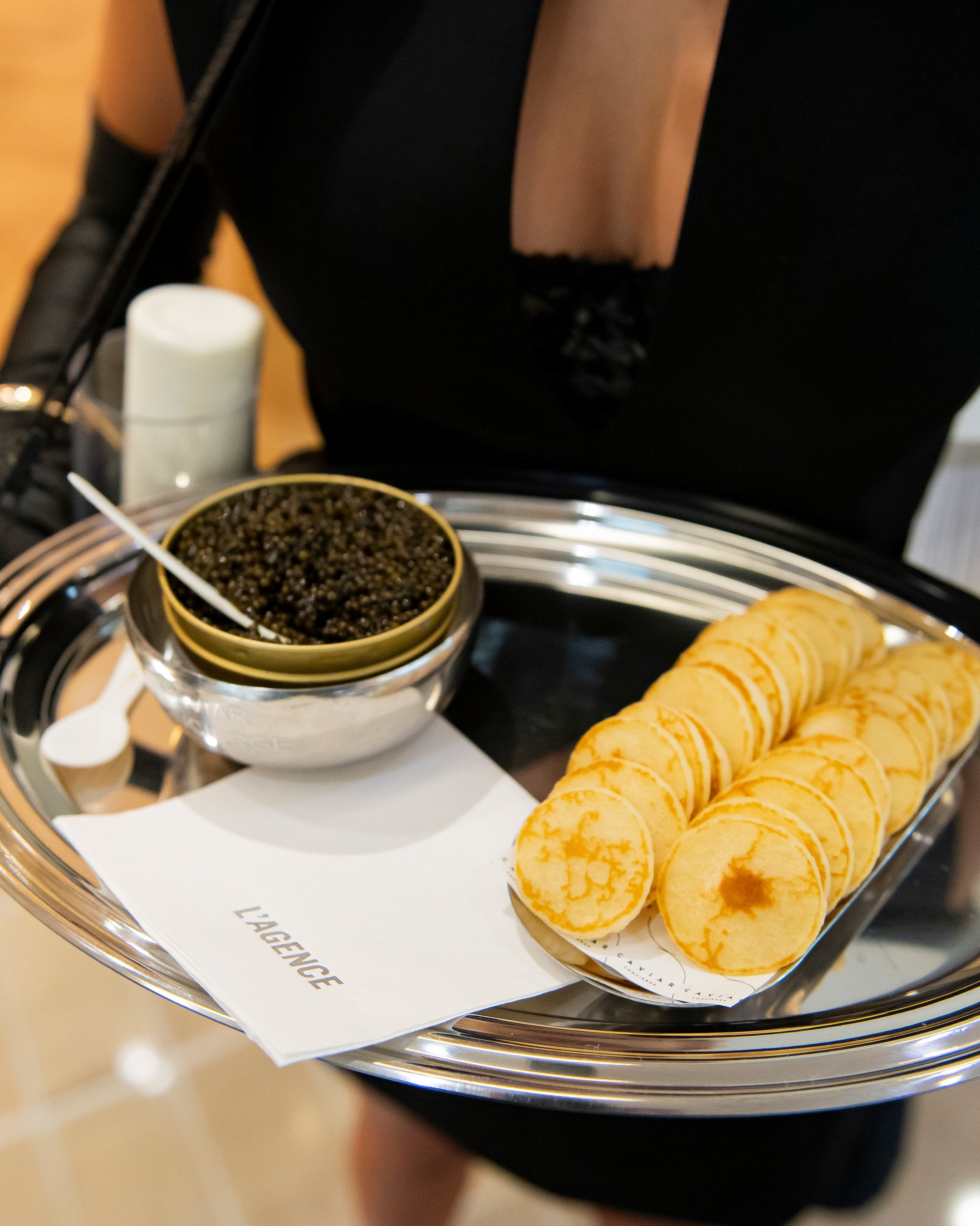 A server in a black dress holds a silver tray with a bowl of caviar and a stack of blini.
