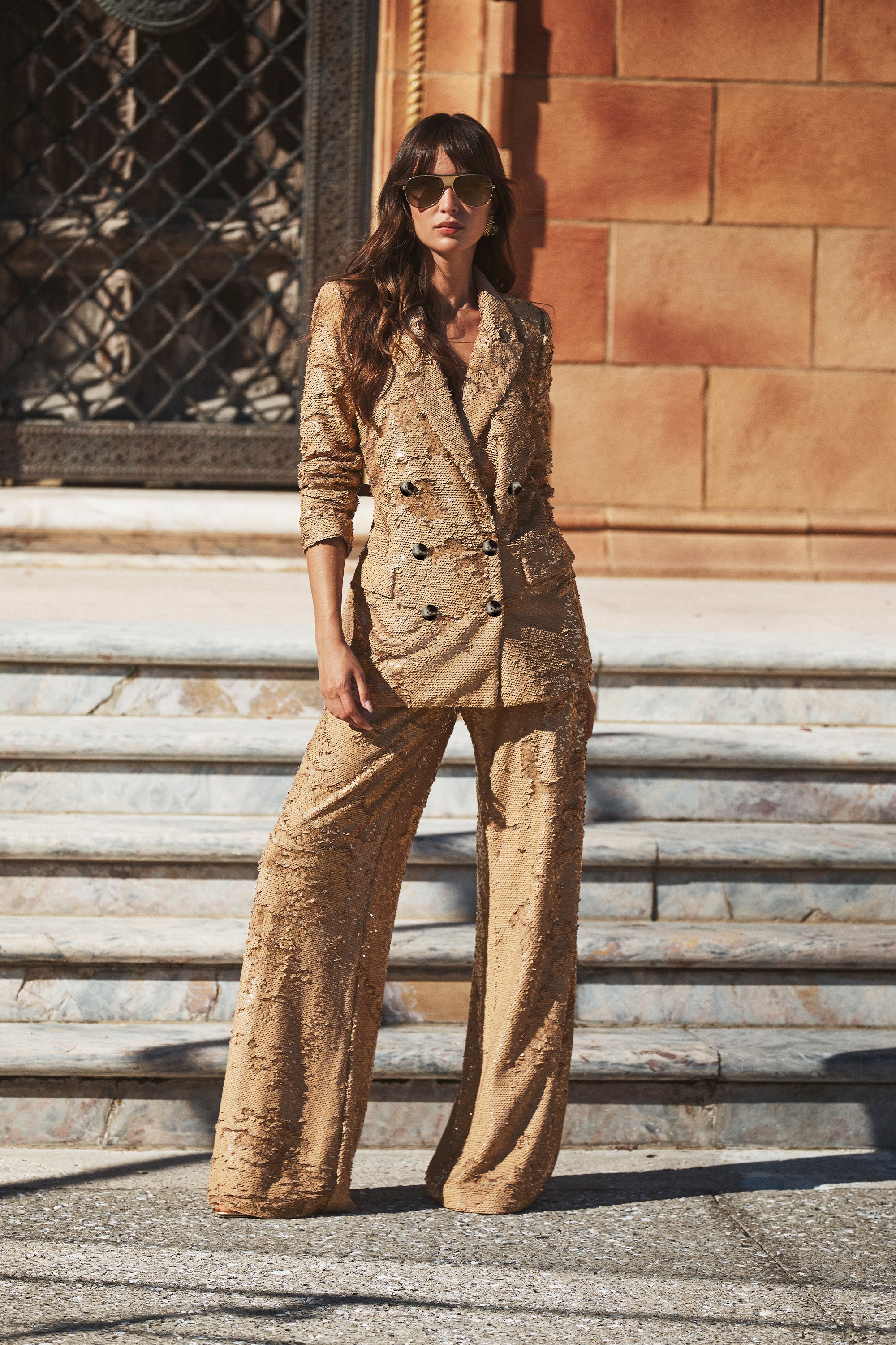 A woman wearing a shiny, gold textured pantsuit and sunglasses stands confidently on marble steps in front of a building with reddish-brown stone walls and a decorative iron window.