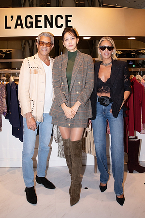 Three stylish adults pose together in front of a L’AGENCE sign in a clothing store. The person in the center wears a plaid blazer and tall boots, while the others wear sunglasses, denim, and fashionable jackets.