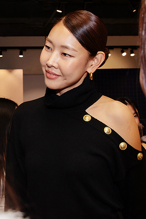 A woman with straight, dark hair styled in a low bun wears a black off-shoulder top with gold buttons, gold hoop earrings, and smiles slightly while standing indoors with people in the background.