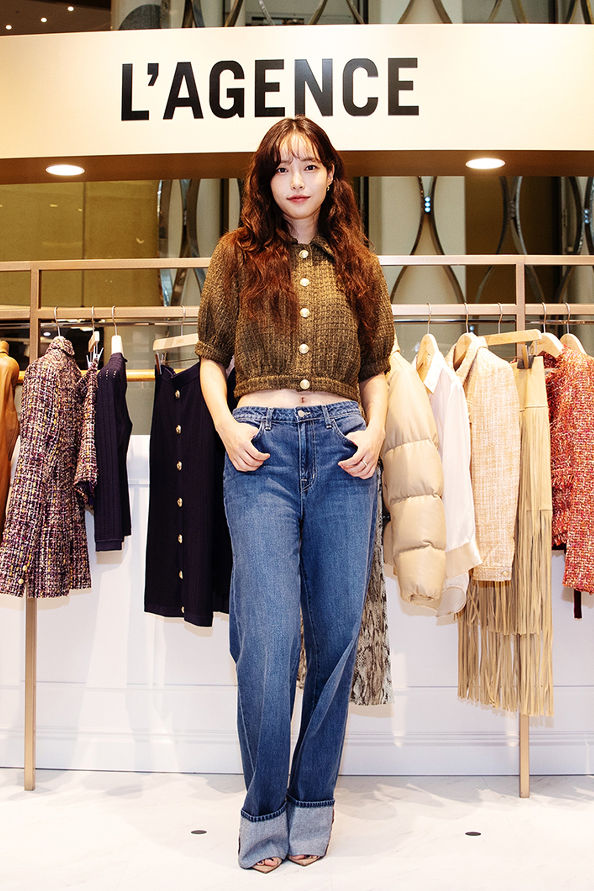 A woman with long, wavy brown hair poses in front of a L’AGENCE store display, wearing a brown knit top and wide-leg jeans, surrounded by hanging clothes in various colors and textures.