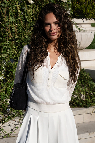A woman with long wavy hair wearing a white top and skirt stands outdoors in sunlight, with greenery and stone steps in the background.