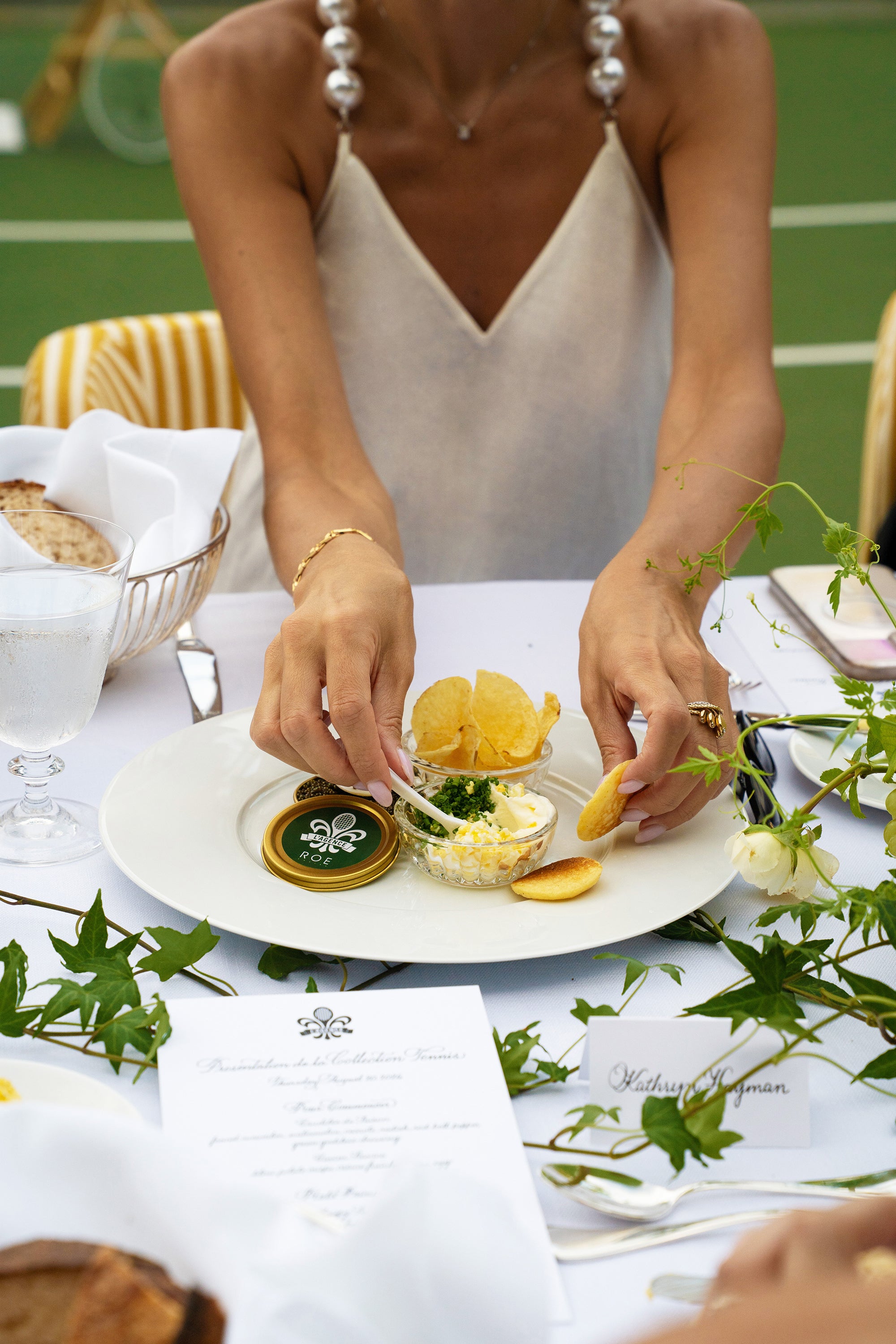 A woman in a white dress sits at an outdoor table, reaching for a dish with potato chips, dip, and herbs. The table is set with bread, a glass of water, and a menu among green leafy decorations.