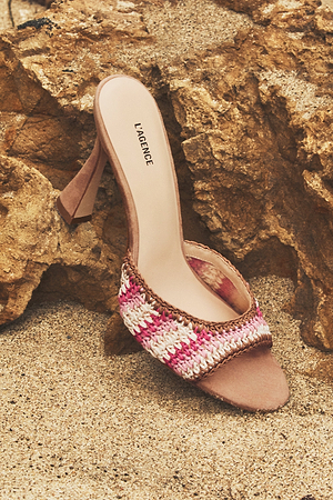 A single heeled sandal with a woven pink, white, and beige strap, and the brand name L’AGENCE on the insole, rests on sandy ground beside a textured rock.
