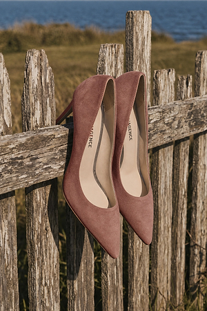 A pair of pink suede pointed-toe high heels are placed on the top of a rustic wooden fence with grass and water visible in the background.