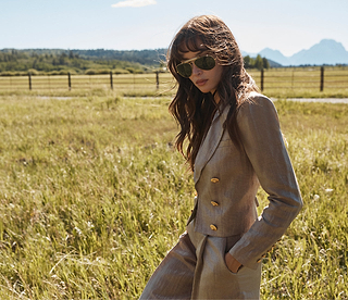 Una donna in tailleur beige e occhiali da sole è in piedi in un prato in una giornata soleggiata, con le montagne e una recinzione visibili sullo sfondo.