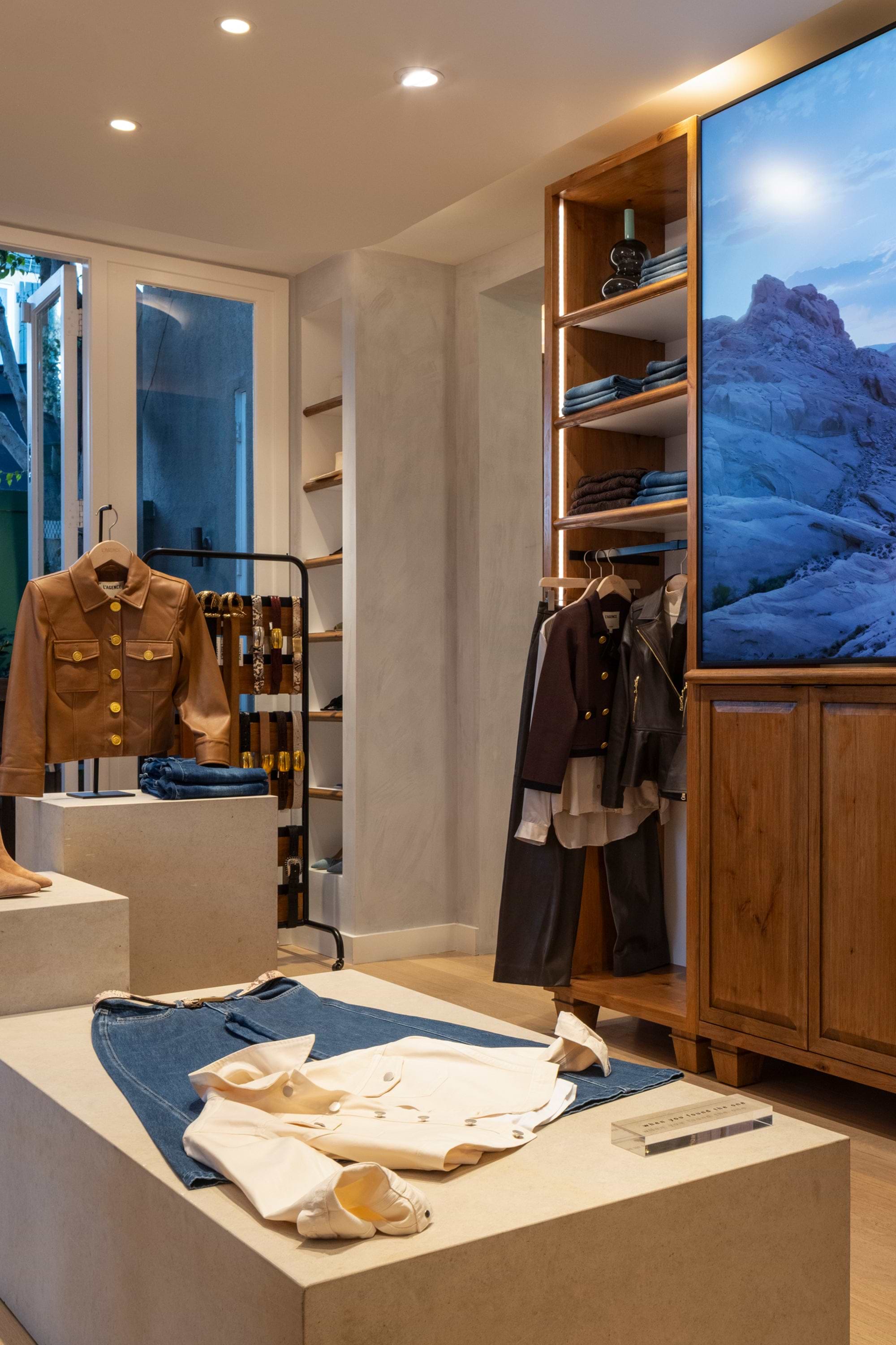 A stylish clothing store interior featuring neatly displayed jackets, shirts, and pants on pedestals and racks, with wooden shelves and a large screen showing a scenic landscape. Warm lighting enhances the inviting atmosphere.