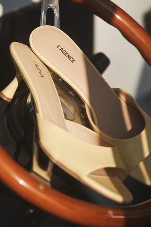 A pair of light yellow heeled mules with L’AGENCE inscribed on the insoles, placed on a wooden and metal steering wheel with soft, natural lighting.