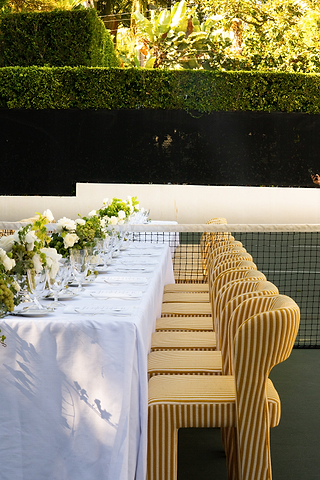 A long table with a white tablecloth, set with plates and flowers, is surrounded by yellow-striped chairs outdoors near a hedge and tennis net.