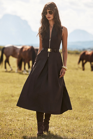 A woman in a sleeveless dark dress with a belt, sunglasses, and boots stands in a grassy field with horses grazing in the background under a partly cloudy sky.