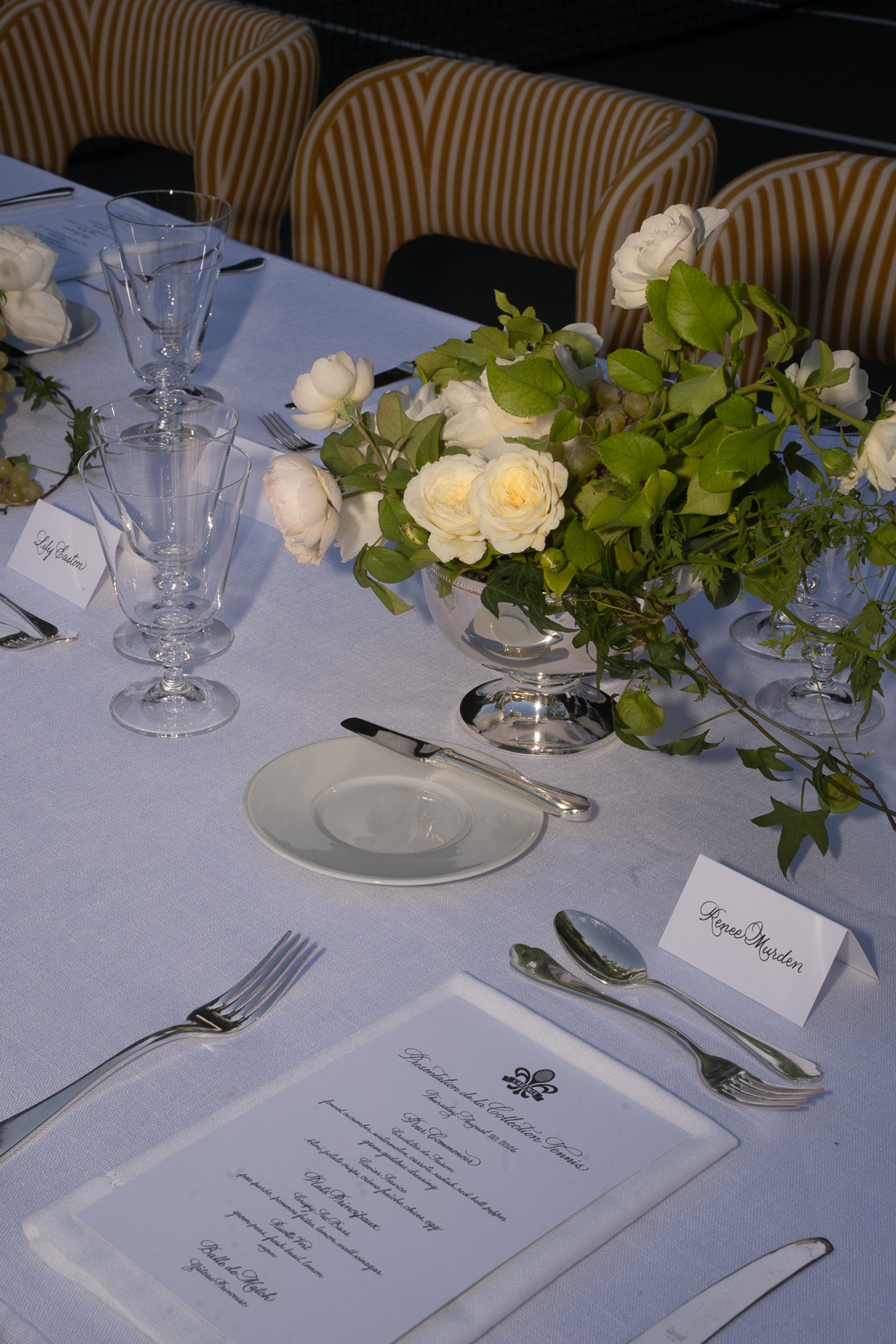 Elegant outdoor table setting with white tablecloth, glassware, silverware, white roses in a vase, name cards, and a printed menu. Striped chairs are in the background. The atmosphere is formal and inviting.