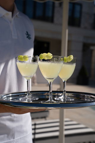 A person in a white shirt holds a silver tray with three stemmed glasses of a pale yellow drink, each garnished with a twisted lemon peel.