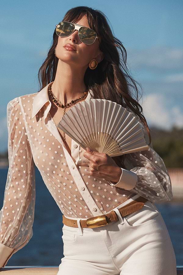 Woman in white sheer polka dot blouse holding a fan with a scenic background.