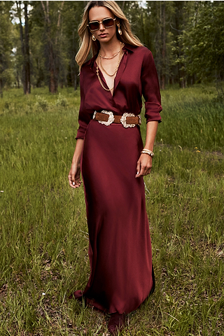 Woman standing outdoors on grass, wearing a long burgundy dress with a deep neckline, gold jewelry, sunglasses, and a tan belt with western-style buckles. Trees and greenery in the background.