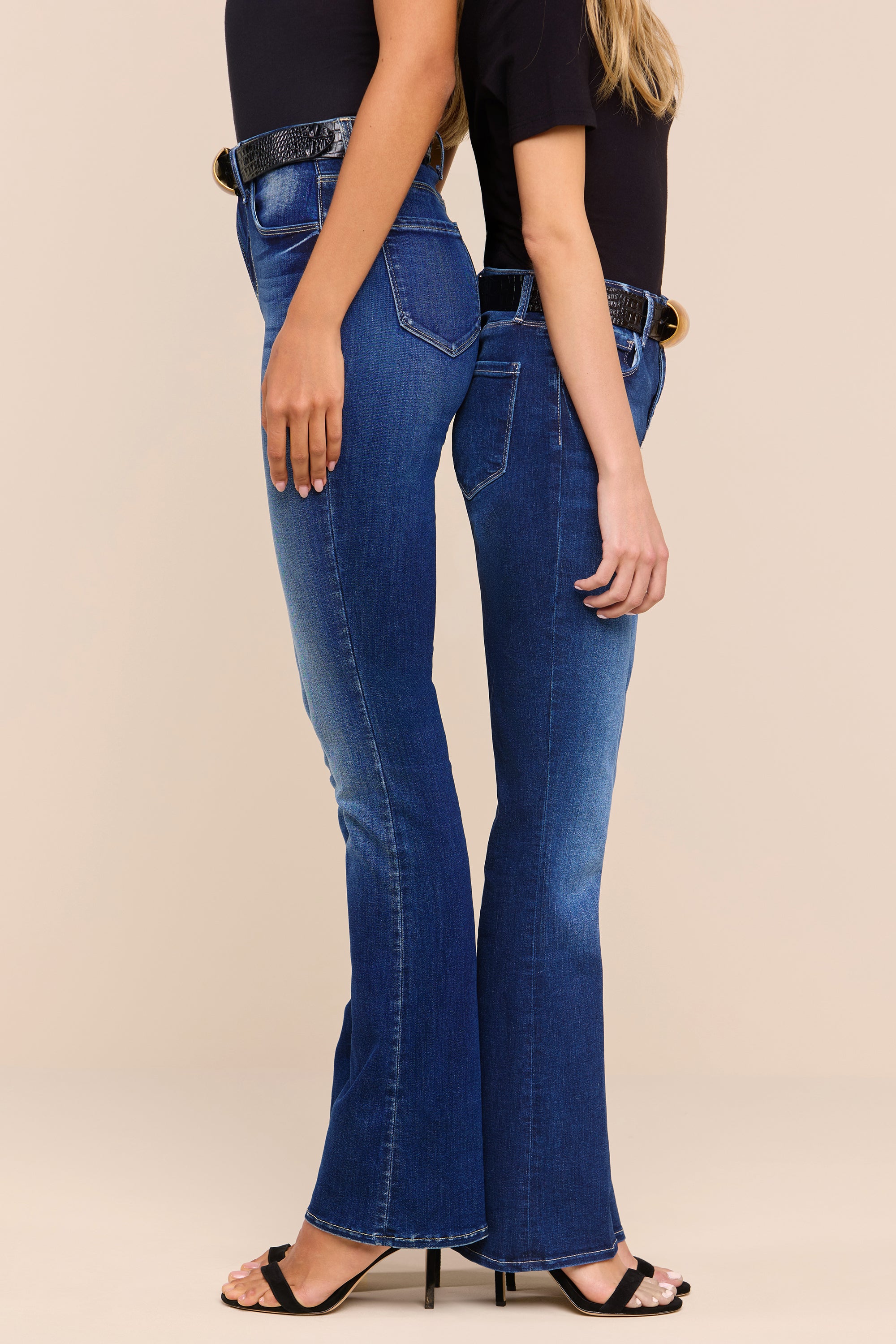 Two women standing side by side, facing opposite directions, wearing dark blue bootcut jeans, black tops, black belts, and open-toed heels against a neutral background.