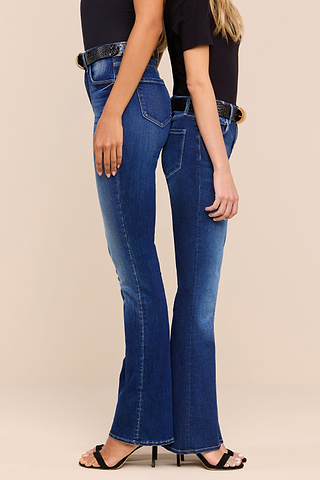 Two women standing side by side, facing opposite directions, wearing dark blue bootcut jeans, black tops, black belts, and open-toed heels against a neutral background.