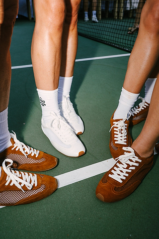 Four people stand in a circle on a green court, wearing white socks and retro sneakers—three in brown, one in white. A tennis net is visible in the background.