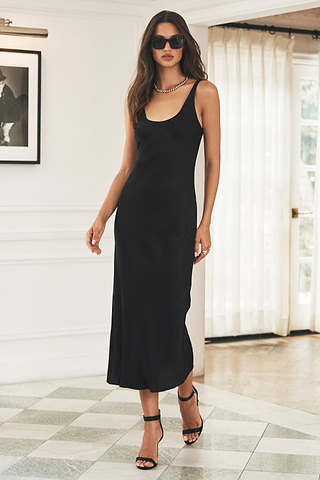 Woman wearing a sleeveless black dress, black heels, sunglasses, and a necklace, standing indoors on a checkered floor with white walls and sheer curtains.