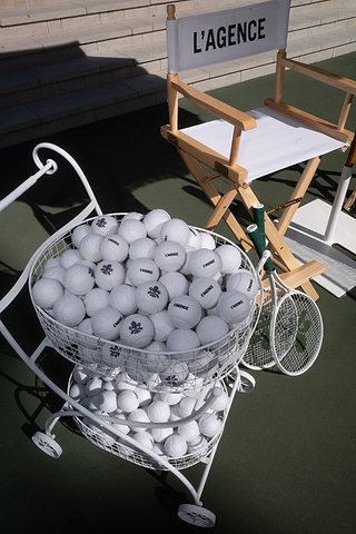 A white cart filled with white tennis balls, two white tennis rackets, and a directors chair labeled LAGENCE on a green tennis court. Sunlight casts shadows on the scene.