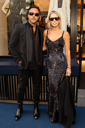 A man in a black suit and sunglasses stands next to a woman in a sleeveless, sequined black gown and sunglasses. They pose together outdoors, with a storefront window behind them.